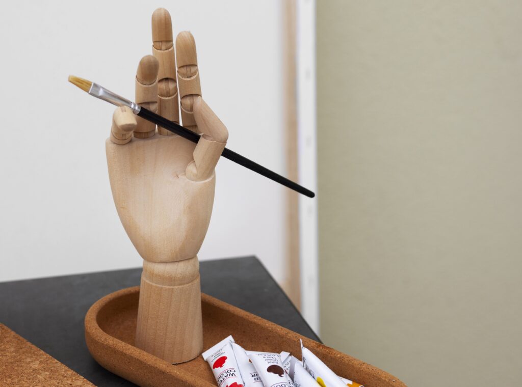 Mão de madeira segurando pincel artístico com tubos de tinta ao lado, representando atividades de arte e pintura na Fredericton Christian Academy.