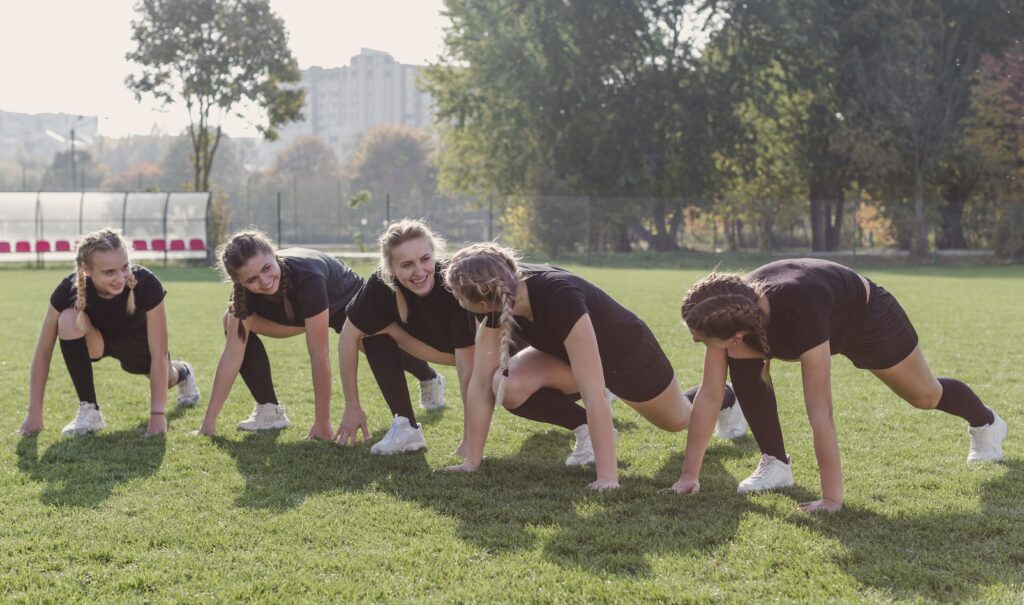 Cinco garotas se alongando em um campo aberto antes do treino de atletismo na Fredericton Christian Academy.