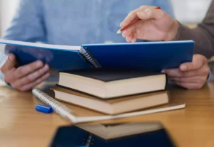 Duas pessoas analisando material acadêmico com livros empilhados sobre a mesa, ilustrando o conceito de Créditos Universitários.