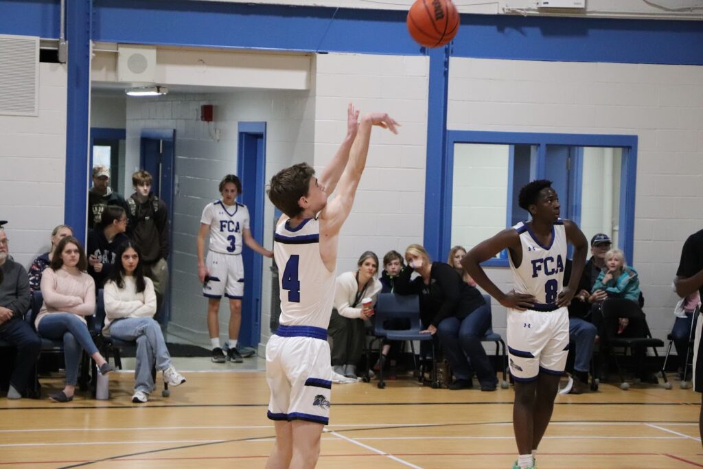 Estudantes da Fredericton Christian Academy jogando basquete com uniforme FCA Eagles em quadra interna, com público assistindo.