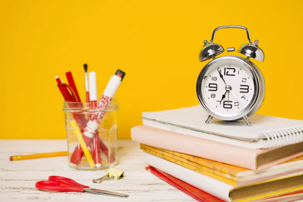 Analog clock on a pile of notebooks with school supplies on the side, symbolizing agility in the registration process.