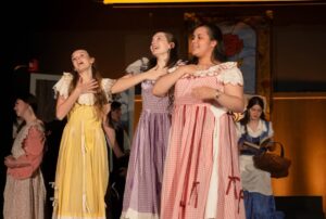 Estudantes cantando caracterizadas como personagens de conto de fadas durante apresentação musical escolar, evidenciando talento vocal e expressão artística.