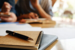 Caderno com espiral e caneta sobre mesa, ao fundo pessoas estudando, simbolizando programas acadêmicos.