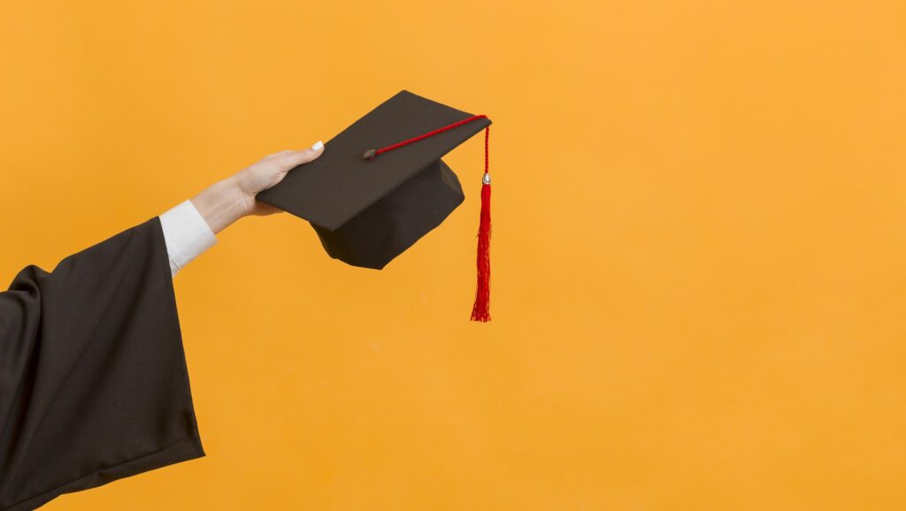 Person in gown holding graduation cap with orange background, representing the academic programs offered by St. Thomas University in Canada.