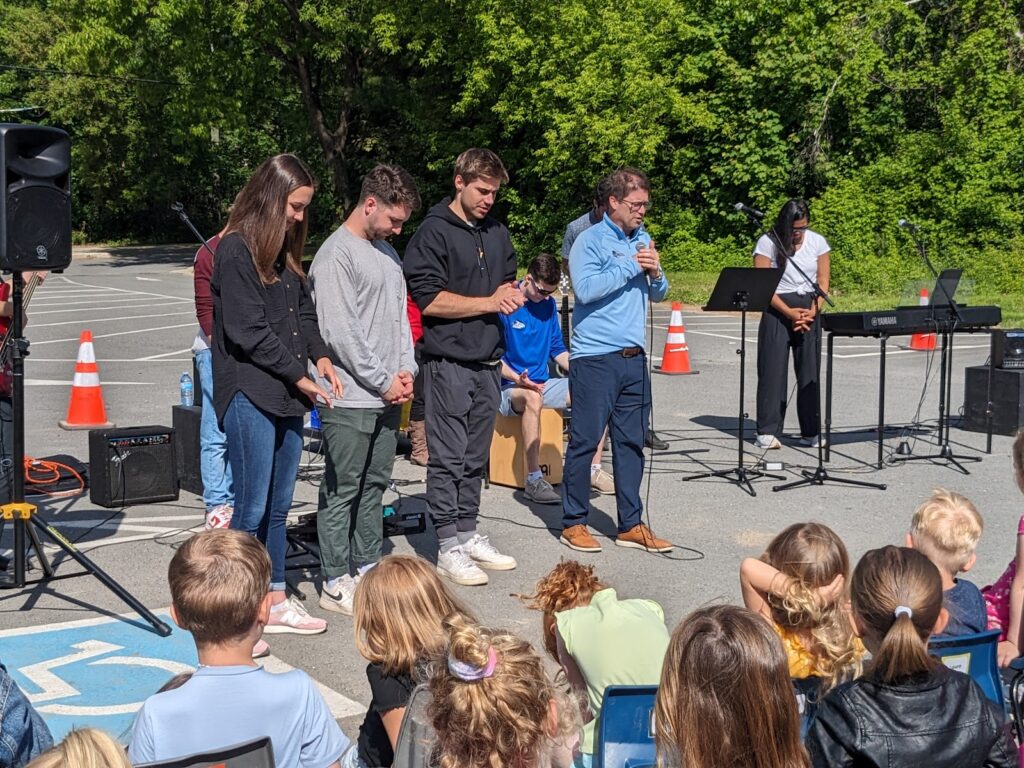 Grupo de alunos e líderes da Fredericton Christian Academy reunidos em oração durante atividade ao ar livre, destacando a vivência da comunidade cristã.