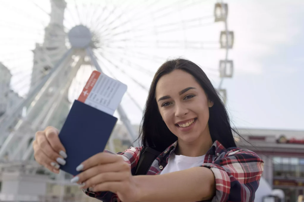 Estudante sorridente segurando passaporte com passagem aérea em frente a roda-gigante, simbolizando viagem para intercâmbio no Canadá.