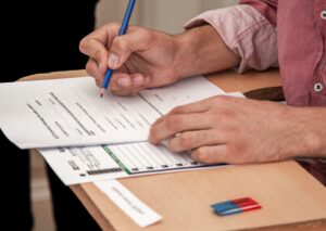 Pessoa preenchendo formulário do teste GMAT com lápis azul sobre carteira escolar.