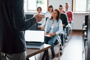 Professor conduzindo aula com grupo de universitários atentos e sorridentes em sala de aula moderna.