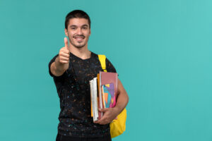Estudante college sorrindo, com mochila amarela e livros nos braços, mostrando sinal de positivo em frente a fundo azul.