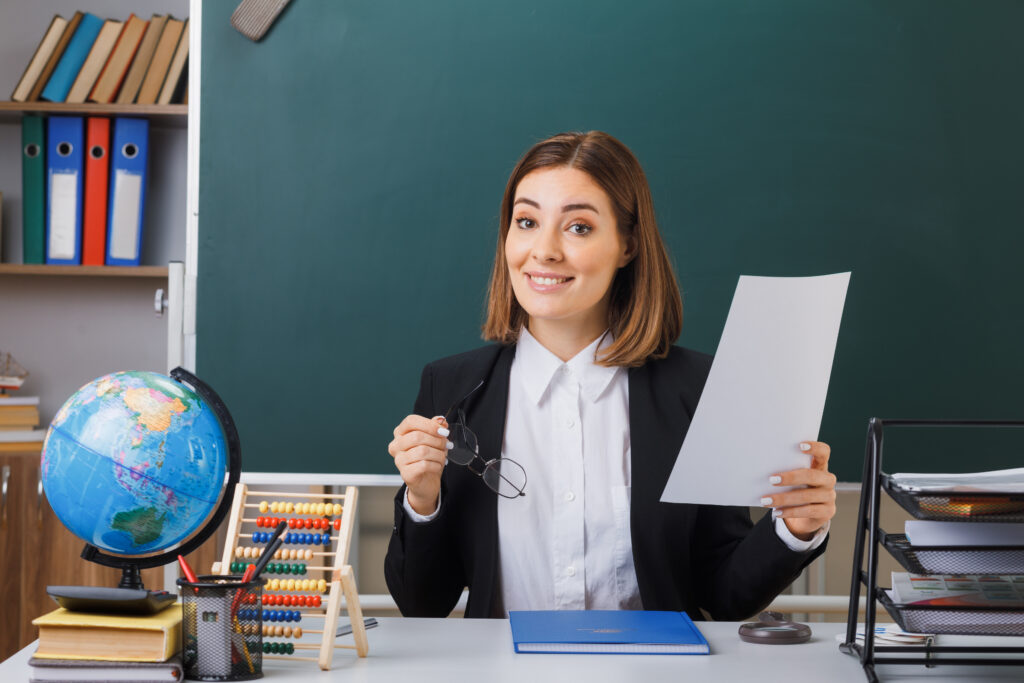 Smiling teacher in classroom with teaching materials around Title: Qualified Teachers Prepared to Teach